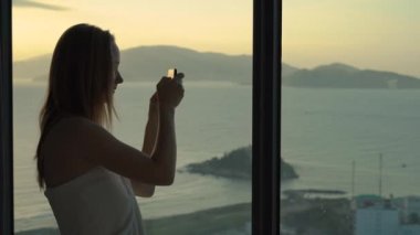 Sahilde ve gündoğumunda panoramik manzaralı bir dairede genç bir kadın. Güzel bir gün doğumunun fotoğrafını çeker.