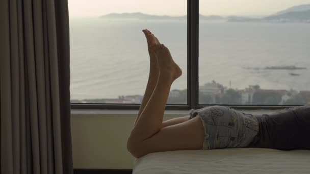 若い女性がベッドの上に横たわり、彼女の後ろにタブレットを使用して海岸にパノラマビューと上昇する太陽の窓があります — ストック動画