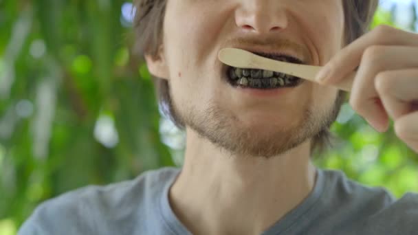 Młody człowiek w zielonym środowisku myje zęby czarnym, aktywnym proszkiem do wybielania zębów. Używa szczoteczki do zębów zrobionej z bambusa. Koncepcja ekologicznego zero odpadów produktów bambusowych — Wideo stockowe