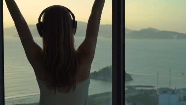 A young woman in an apartment with a panoramic view on the coast and sunrise. She puts on wireless earphones and listens to the music — Stock Video