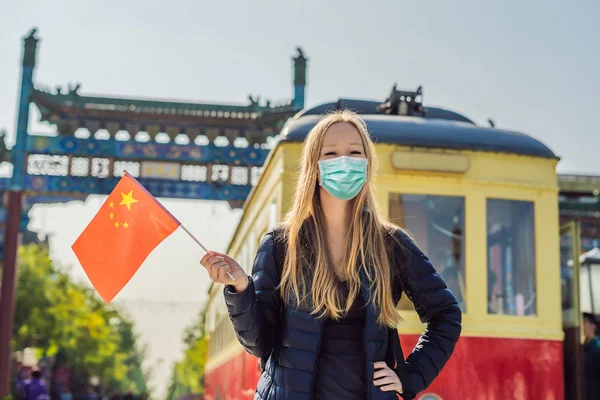 Enjoying vacation in China. Young woman in medical mask with national chinese flag on the background of the old Chinese street. Travel to China concept. Tourists fear the 2019-ncov virus. Medical