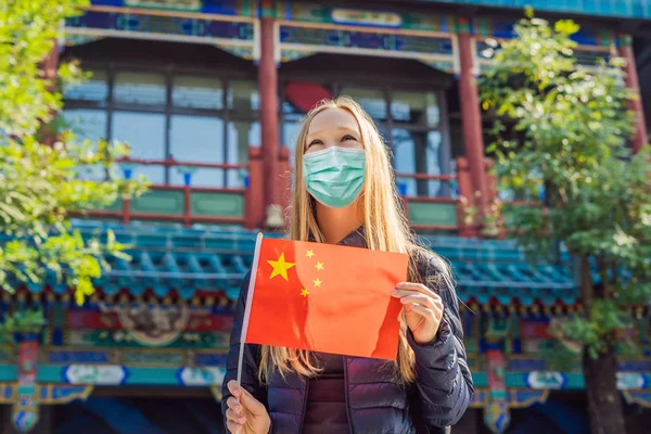 Enjoying vacation in China. Young woman in medical mask with national chinese flag on the background of the old Chinese street. Travel to China concept. Tourists fear the 2019-ncov virus. Medical