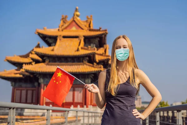 Enjoying vacation in China. Young woman in medical mask with national chinese flag in Forbidden City. Travel to China concept. Tourists fear the 2019-ncov virus. Medical masked tourists