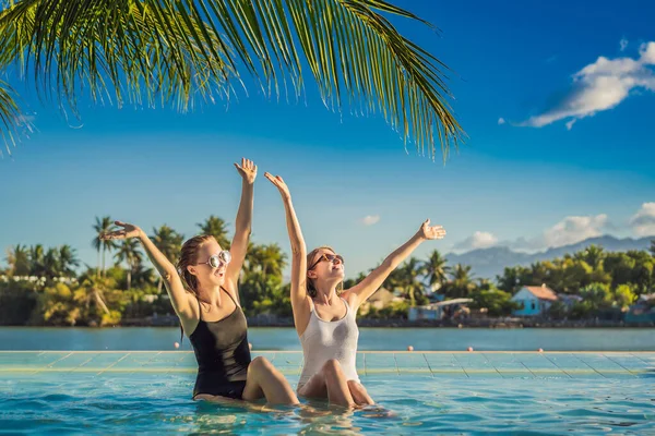 Deux belles jeunes femmes assises au bord de la piscine d'une station balnéaire pendant les vacances d'été. Fête, tourisme dans les pays tropicaux — Photo