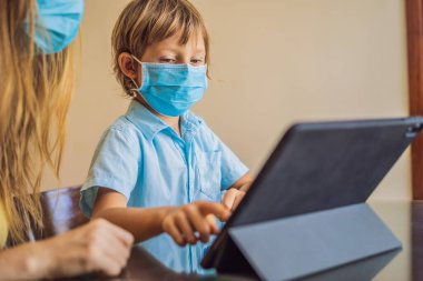 Çocuk evde bir tablet kullanarak online çalışıyor. Annem öğrenmesine yardım ediyor. Koronovirüsten korunmak için maskeli anne ve oğul. Karantina sırasında ders çalışmak. Küresel salgın covid19 virüsü