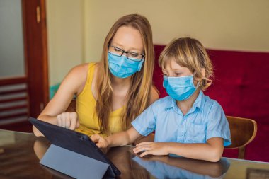 Çocuk evde bir tablet kullanarak online çalışıyor. Annem öğrenmesine yardım ediyor. Koronovirüsten korunmak için maskeli anne ve oğul. Karantina sırasında ders çalışmak. Küresel salgın covid19 virüsü