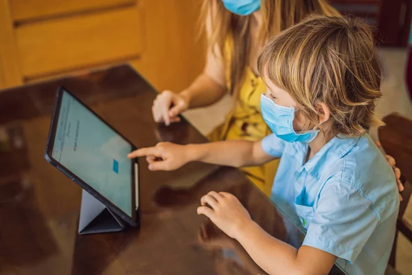 Boy studying online at home using a tablet. Mom helps him learn. Mom and son in medical masks to protect against coronovirus. Studying during quarantine. Global pandemic covid19 virus