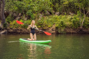 Kalk ayağa kürek çeken kadın gölde kürek çekiyor mavi suda kürek çekerken mutlu duruyor. Genç Kadın Kürek Çekme Kurulu 'nda