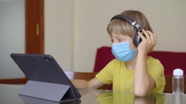 Koruyucu maskeli küçük bir çocuk karantina sırasında evde oturur ve internet okulunda tablet kullanarak ders çalışır. Corona virüsü konsepti