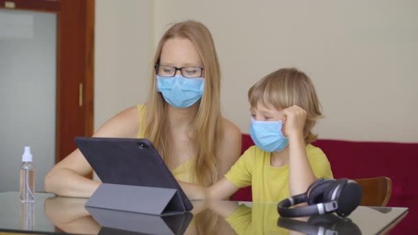 Una mujer joven y su pequeño hijo se sientan en una casa durante la cuarentena y estudian en la escuela de Internet usando una tableta. Concepto de virus Corona — Vídeo de stock