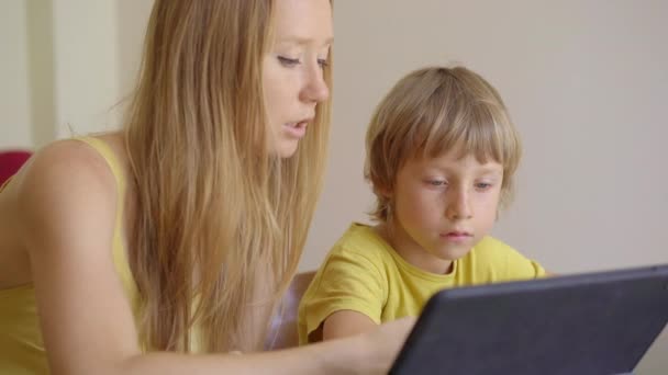 Una mujer joven y su pequeño hijo se sientan en una casa durante la cuarentena y estudian en la escuela de Internet usando una tableta. Concepto de virus Corona — Vídeo de stock