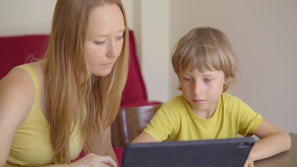Una mujer joven y su pequeño hijo se sientan en una casa durante la cuarentena y estudian en la escuela de Internet usando una tableta. Concepto de virus Corona — Vídeo de stock