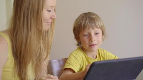 Una mujer joven y su pequeño hijo se sientan en una casa durante la cuarentena y estudian en la escuela de Internet usando una tableta. Concepto de virus Corona — Vídeos de Stock