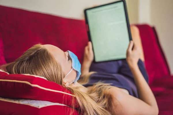 Woman in medical mask reads a book on the couch. Home quarantine. Stay at home. Coronovirus