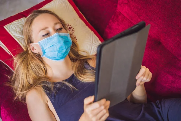 Kobieta w masce medycznej czyta książkę na kanapie. Kwarantanna domowa. Zostań w domu. Koronowirus — Zdjęcie stockowe