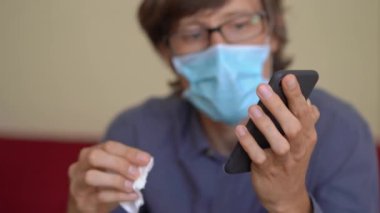 Yüz maskesi takan genç bir adam, koronavirüs kendini izole ederken evden çalışır. Telefonunu dezenfekte etmek için alkol dezenfektanı kullanıyor.