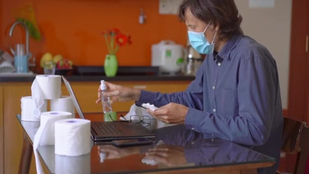 A young man wearing a face mask sitting at home during coronavirus self-isolation. He uses an alcohol sanitizer to disinfect everything that surrounds him. Slowmotion shot — Stock Video