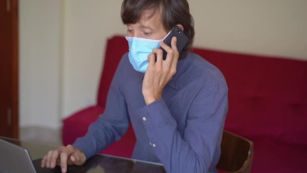 Un giovane uomo che indossa una maschera facciale lavora da casa durante l'auto-isolamento coronavirus. Parla con un cellulare. Colpo di rallentamento . — Video Stock