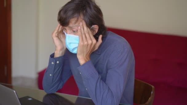 Een jongeman met een gezichtsmasker werkt vanuit huis tijdens de zelfisolatie van het coronavirus. Hij is het beu om thuis te blijven en bezorgd te zijn over de toekomst. Langzame schot — Stockvideo