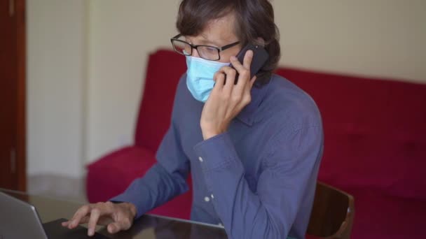 Ein junger Mann mit Gesichtsmaske arbeitet von zu Hause aus während der Selbstisolation mit dem Coronavirus. Er telefoniert mit einem Handy. Zeitlupenschuss. — Stockvideo