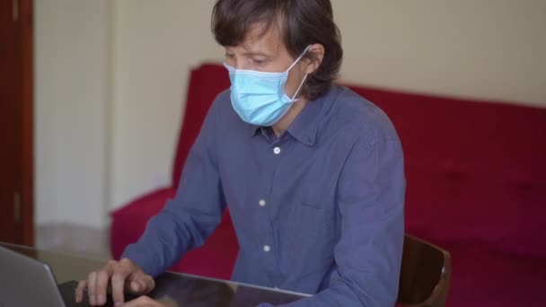 Een jongeman met een gezichtsmasker werkt vanuit huis tijdens de zelfisolatie van het coronavirus. Hij praat met een mobieltje. Langzame schot. — Stockvideo