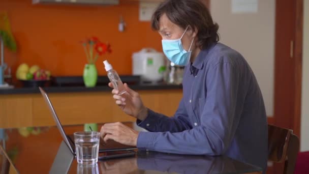 Un giovane uomo che indossa una maschera facciale parla con qualcuno attraverso una videoconferenza da casa durante l'auto-isolamento coronavirus. Si applica disinfettante per le mani sulle mani e lo mostra al suo amico — Video Stock