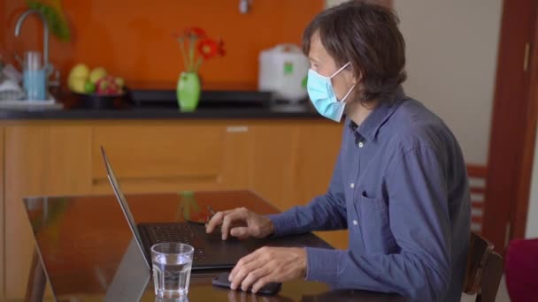 Een jongeman met een gezichtsmasker werkt vanuit huis tijdens de zelfisolatie van het coronavirus. Hij praat met een mobieltje. Langzame schot. — Stockvideo