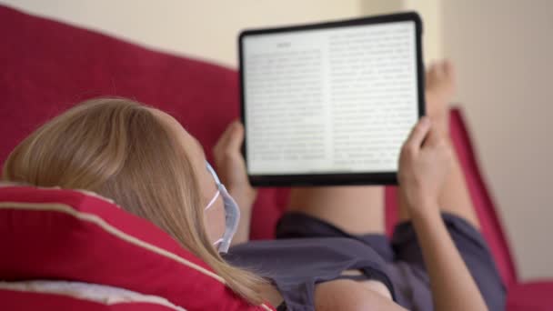 En ung kvinna i medicinsk ansiktsmask lägger sig på en soffa och läser en elektronisk bok. Hon stannar hemma under coronavirus självisolering — Stockvideo