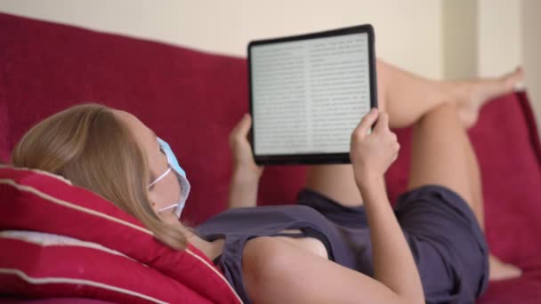 Uma jovem mulher em uma máscara facial médica coloca em um sofá e lê um livro eletrônico. Ela fica em casa durante o auto-isolamento do coronavírus — Vídeo de Stock