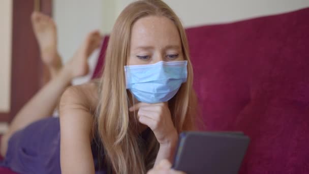 Een jonge vrouw met een medisch gezichtsmasker ligt op de bank en leest een elektronisch boek. Ze blijft thuis tijdens zelfisolatie van het coronavirus. — Stockvideo