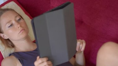 Genç bir kadın kanepeye uzanıp elektronik kitap okuyor. Koronavirüs kendini izole ederken evde kalıyor.