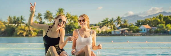Deux belles jeunes femmes assises au bord de la piscine d'une station balnéaire pendant les vacances d'été. Fête, tourisme dans les pays tropicaux BANNER, LONG FORMAT — Photo