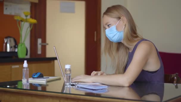 Una mujer joven con una máscara facial médica trabaja desde casa durante el autoaislamiento del coronavirus. Trabajo remoto concepto — Vídeo de stock