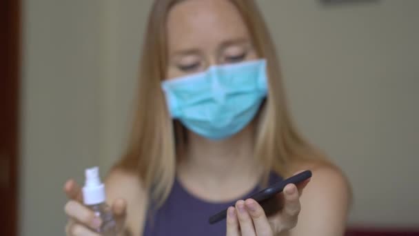 Mladá žena s obličejovou maskou pracuje z domova během sebeizolace koronaviru. Používá dezinfekci na alkohol, aby si vydezinfikovala telefon. — Stock video