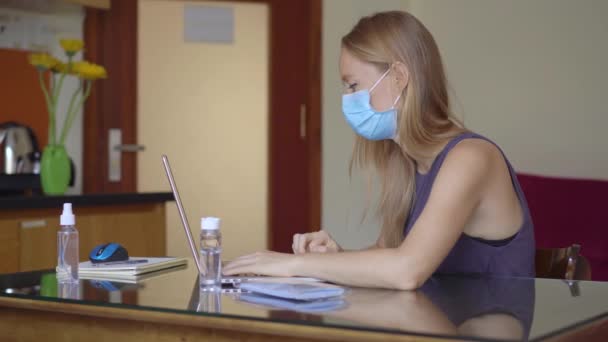 En ung kvinna som bär en medicinsk ansiktsmask arbetar hemifrån under coronavirus självisolering. Fjärrstyrt arbete — Stockvideo