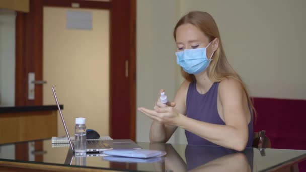 Een jonge vrouw met een medisch gezichtsmasker werkt vanuit huis tijdens de zelfisolatie van het coronavirus. Ze brengt handreiniger aan op haar handen. — Stockvideo