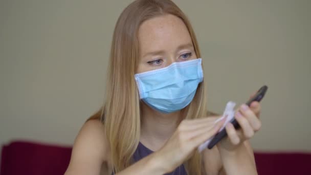 Una mujer joven con una máscara facial trabaja desde casa durante el autoaislamiento del coronavirus. Usa un desinfectante de alcohol para desinfectar su teléfono. — Vídeos de Stock