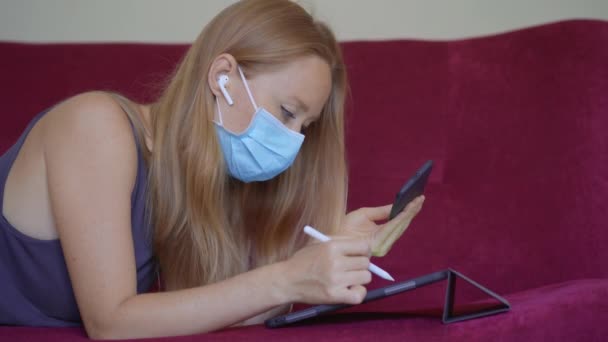 Een jonge vrouw met een medisch gezichtsmasker werkt vanuit huis tijdens de zelfisolatie van het coronavirus. Zelfontplooiing tijdens sociaal isolement. Een vrouw trekt op een tablet. Werken op afstand concept — Stockvideo