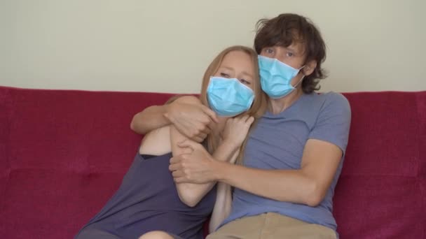 A young man and woman during self-isolation sit at home on a couch. The man hugs his scared wife. They wear medical face masks — Stock Video