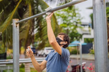 Adam spordan önce yatay barı dezenfekte eder. Coronavirus salgını sırasında tıbbi maskeli bir adam spor yapıyor.