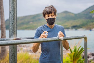 Adam spordan önce yatay barı dezenfekte eder. Coronavirus salgını sırasında tıbbi maskeli bir adam spor yapıyor.