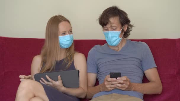 Un hombre y una mujer jóvenes durante el autoaislamiento se sientan en casa en un sofá. Tablet no reconoce a la mujer con una máscara médica — Vídeos de Stock