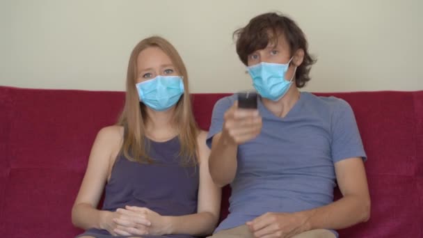 A young man and woman during self-isolation sit at home on a couch looking at the TV. They wear medical face masks — Stock Video