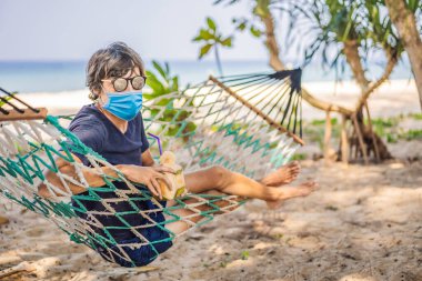Plajda bir hamakta maske takmış genç bir adam ve bir içki turistleri 2019-Ncov virüsünden korkuyorlar. Tıbbi maskeli turistler