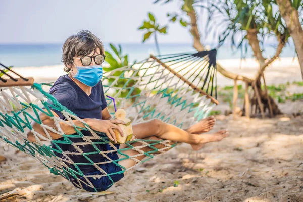 Hombre joven con máscara médica en la playa en una hamaca con una bebida Los turistas temen el virus 2019-ncov. Turistas médicos enmascarados —  Fotos de Stock
