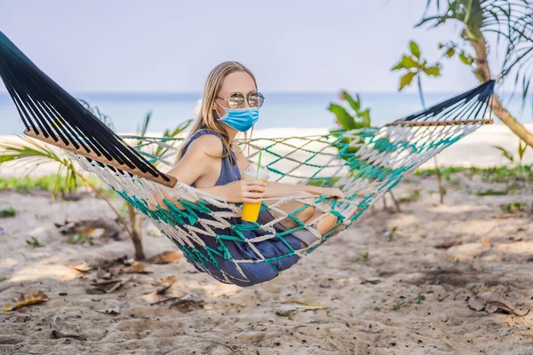 Mujer joven con máscara médica en la playa en una hamaca con una bebida Los turistas temen el virus 2019-ncov. Turistas médicos enmascarados — Foto de Stock