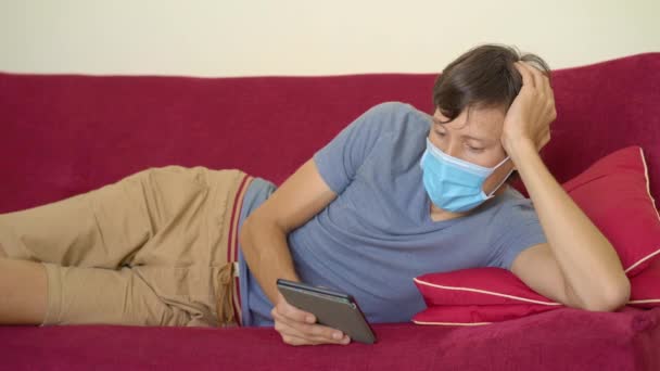 E-book en online onderwijs. Een man leest thuis een elektronisch boek tijdens quarantaine. Een man met een medisch masker bleef thuis in zelfisolatie. — Stockvideo