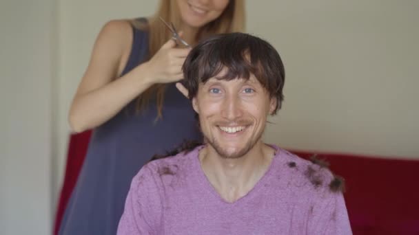 Uma jovem faz cortes de cabelo para o namorado ou marido por causa do confinamento. Ela está tentando seguir instruções de vídeo. O homem tem medo que o corte de cabelo dele seja horrível. Conceito de isolamento social — Vídeo de Stock