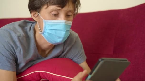 E-book en online onderwijs. Een man leest thuis een elektronisch boek tijdens quarantaine. Een man met een medisch masker bleef thuis in zelfisolatie. Uiteindelijk valt hij in slaap.. — Stockvideo