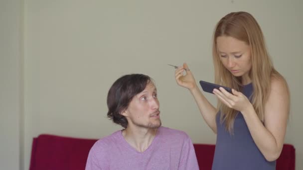 Una giovane donna fa tagli di capelli al suo fidanzato o marito a causa di isolamento. Sta cercando di seguire le istruzioni video. L'uomo ha paura che il suo taglio di capelli sarà terribile. Il concetto di isolamento sociale — Video Stock